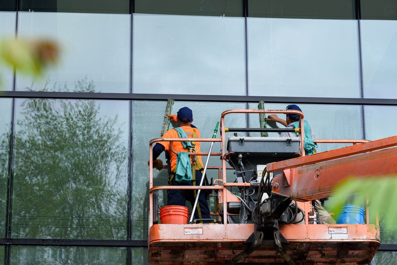 about-01 Two workers cleaning windows on a glass building facade using a lift platform, enhancing building aesthetics.
